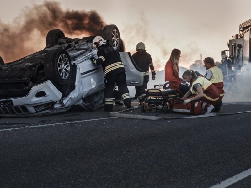 photo of a car accident scene with vehicle fliiped over and woman being treated by paramedics
