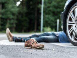 Person lying on a road with one brown shoe in the foreground and a car wheel visible to the right, suggesting an accident scene.