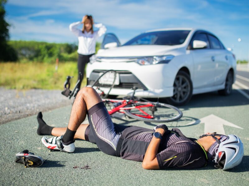 Cyclist lying on the road after a crash, helmet off nearby, bike on its side, white car stopped in the background, bystander distressed.